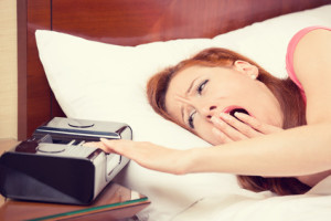 woman extending hand to alarm clock yawning lies in bed