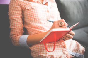 A woman in a check shirt writing in a red diary - it's a great idea to journal about any anxieties you may have when anticipating going on holidat with ME, CFS or Fibromyalgia