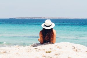 A woman in a straw hat reclines on a white sand beach in front of an azure ocean - going on holiday when you have ME, Chronic Fatigue or Fibromyalgia