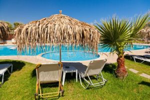 A couple of sunloungers under a palm leaf umbrella beside a turquoise swimming pool - how to ask for what you need when on holiday with ME, CFS or Fibromyalgia